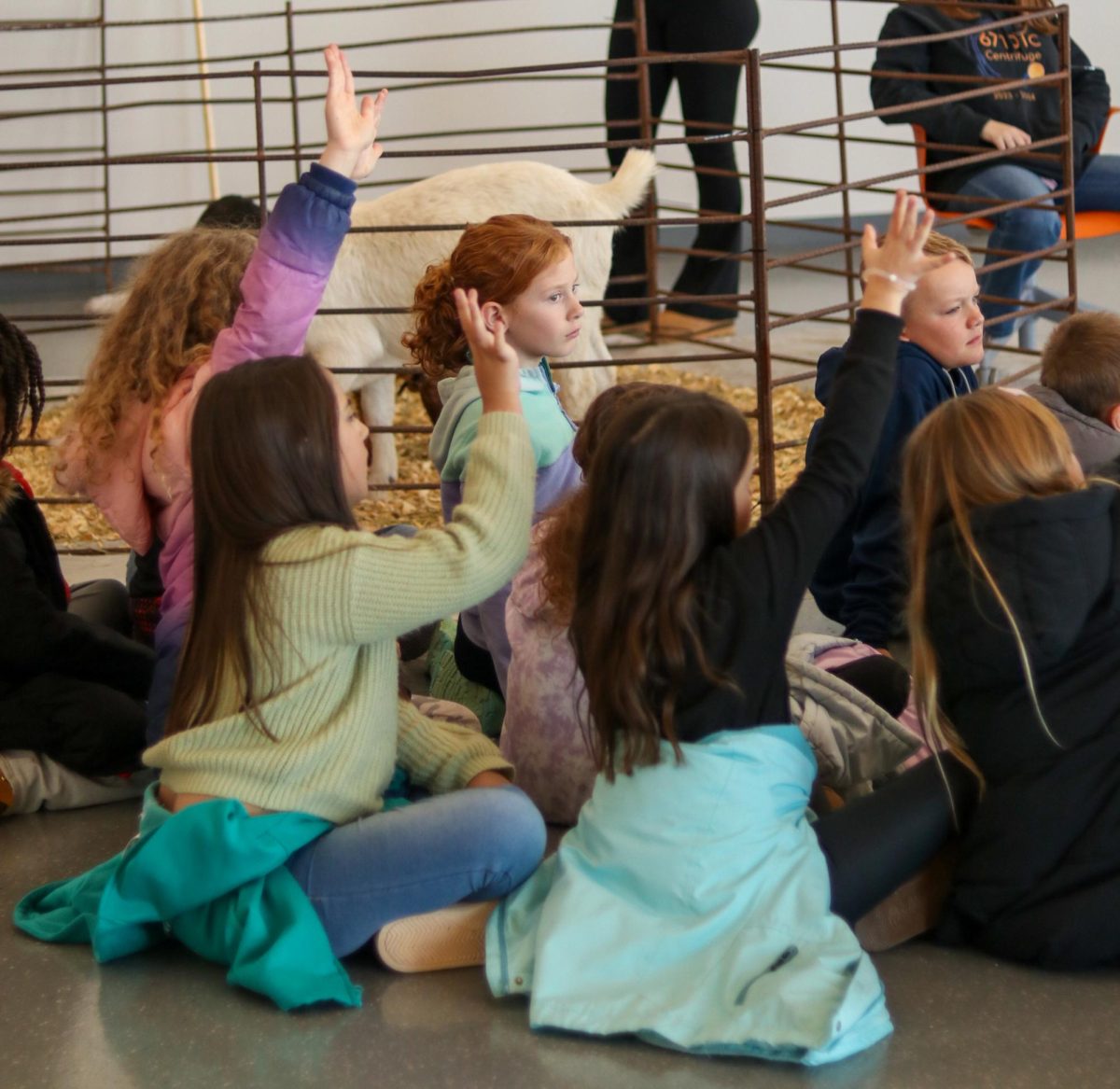 Multiple students, some raising their hands, sitting in a group on the floor.