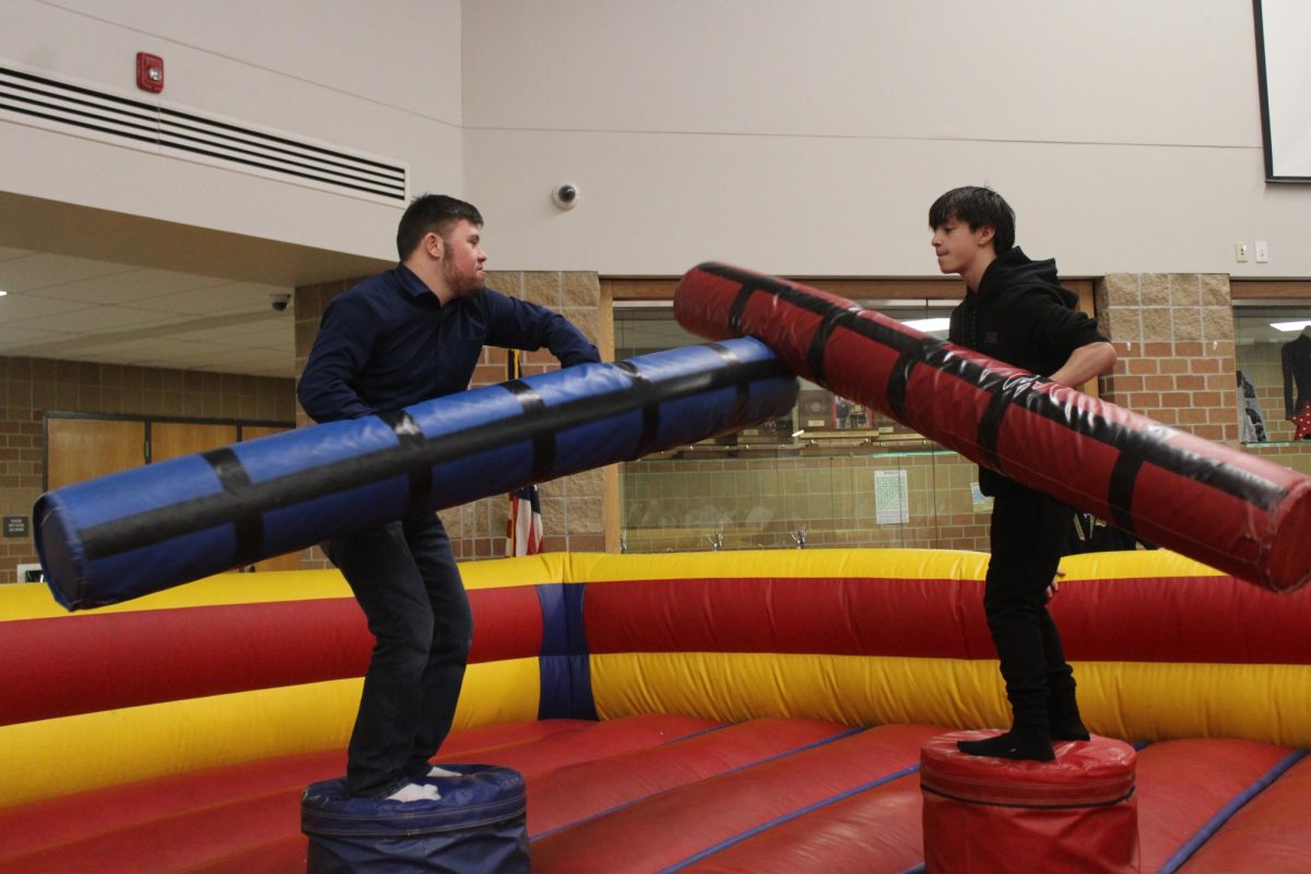 Two teen boys stand on inflatable podiums while holding padded poles to try to knock each down